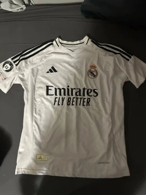 Real Madrid matchtröja Adidas vit - Säljer en vit Real Madrid matchtröja från Adidas med svarta detaljer och klubbmärke på bröstet. Tröjan har svarta ränder på axlarna, trycket 'Emirates Fly Better' och patch på ärmen. Tillverkad i lätt och ventilerande material.