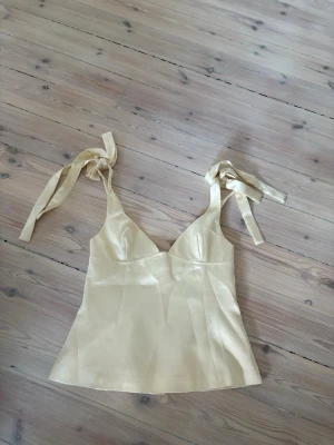 Ljusgul satinlinnetopp med knytband - Säljer en ljusgul linnetopp i satin med breda axelband som knyts i rosetter. Toppen har en lätt utsvängd passform och v-ringad urringning. Perfekt för dig som gillar en feminin och trendig look. Aldrig använd då den inte passar därav bra skick!