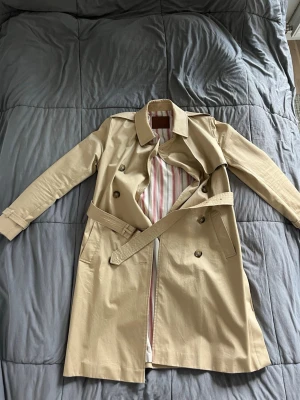 Beige trenchcoat från Massimo Dutti - Stilren beige trenchcoat från Massimo Dutti med klassisk dubbelknäppning, bälte i midjan och snyggt randigt innerfoder i vitt, rött och svart. Perfekt för dig som vill ha en tidlös och trendig jacka till garderoben.