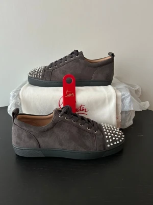 Gråa Louboutin sneakers med nitar - Snygga gråa sneakers från Christian Louboutin i mocka med silverfärgade nitar på tån och klassisk röd sula. Lågt skaft och svarta snören ger en clean look med edgy detaljer. Perfekta för dig som vill sticka ut med exklusiv streetstyle. Allt OG medföljer förutom OG box