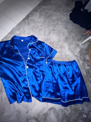 Blå satinpyjamas med korta ärmar - Snygg blå pyjamas i glansigt satinmaterial med kortärmad skjorta och matchande shorts. Skjortan har vita knappar och vit passpoal längs kanterna samt en liten bröstficka. Shortsen har resår i midjan och vit kant nertill. Perfekt för mysiga kvällar hemma.