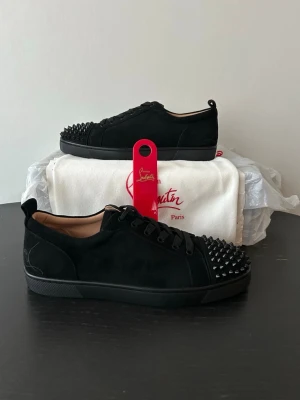Svarta spikes-sneakers Louboutin - Snygga svarta sneakers i mocka från Christian Louboutin med svarta nitar på tån och klassisk röd sula. Lågt skaft och svarta skosnören ger en clean look. Perfekta för dig som vill sticka ut med exklusiv streetstyle. Allt OG medföljer förutom OG box