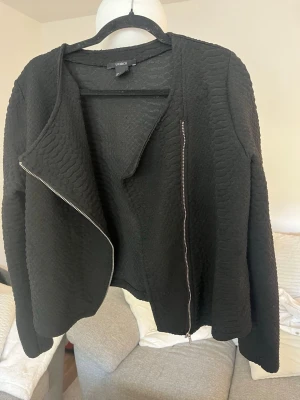 Svart skinnjacka med struktur från Lindex - Snygg svart skinnjacka från Lindex med cool struktur och asymmetrisk dragkedja. Jackan har lång ärm och en modern, lite boxig passform. Perfekt för dig som vill ha en edgy look och gillar detaljer som sticker ut.