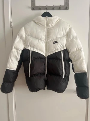 Svartvit pufferjacka från Nike - Säljer en snygg svartvit pufferjacka från Nike med huva och dragkedja framtill. Jackan har två stora fickor med vita dragkedjor och en cool färgblock-design. Perfekt för kalla dagar när du vill hålla stilen och värmen.