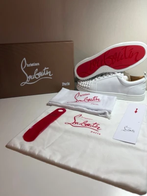 Vita Christian Louboutin sneakers - Snygga vita sneakers från Christian Louboutin med ikonisk röd sula och nitar på tån. Skorna är låga och har en stilren design i läder med vita snören. Perfekta för dig som vill sticka ut med exklusiva detaljer och klassisk streetstyle.