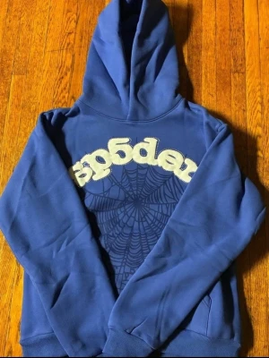 Blå hoodie med spindelnätsmönster - Blå hoodie med stor huva och ett coolt spindelnätsmönster på bröstet. Framtill står det 'sp5der' i vit, fluffig text. Hoodien har ribbade muddar vid ärmar och nederkant. Perfekt för dig som gillar streetwear och vill sticka ut.