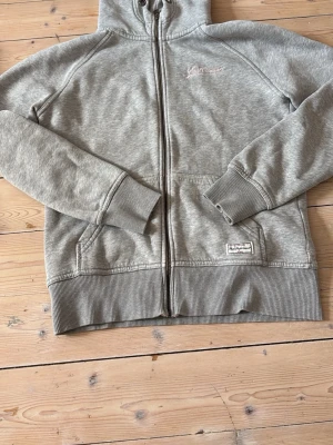 Grå zip hoodie från Peak Performance - Säljer en grå zip hoodie från Peak Performance med broderad logga i ljusrosa på bröstet. Storlek M men ganska liten i storleken Hoodien har huva med dragsko, två fickor fram och ribbade muddar vid ärmar och nederkant. Perfekt för chill dagar eller till träningen.