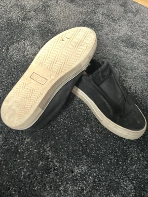 Svarta slip-on sneakers från Arigato - Svarta slip-on sneakers från Arigato med elastiska band över vristen och vit sula. Ovandelen är i mockaliknande material och designen är clean utan snörning. Perfekta för dig som gillar minimalistisk stil och vill ha något enkelt att dra på sig. 