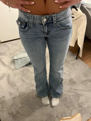 Gina tricot jeans - Säljer ett par ljusblå bootcut jeans med snygga fickdetaljer både fram och bak. Jeansen har låg midja, dubbla knappar i linningen och dekorativa sömmar. Perfekta för en trendig och avslappnad look.