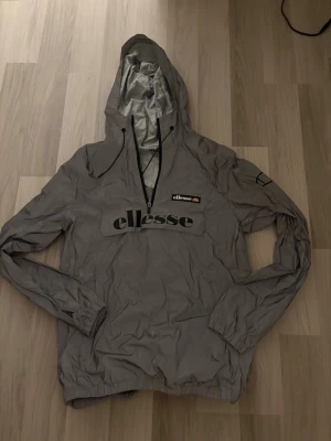 Grå vindjacka från Ellesse med huva - Snygg grå vindjacka från Ellesse med stor logga på bröstet och patch på vänster ärm. Jackan har huva med dragsko, halv dragkedja framtill och elastiska muddar. Perfekt för dig som vill ha en sportig och stilren look.