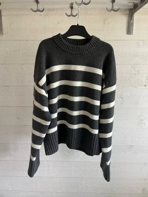 Mörkgrå stickad tröja med ränder H&M - Mysig mörkgrå stickad tröja från H&M med breda vita horisontella ränder. Tröjan har rund halsringning och ribbstickade muddar vid ärmslut och nederkant. Perfekt för lager på lager och chill stil.