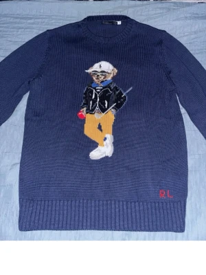 Ralph Lauren Björn tröja Bear - Mörkblå stickad tröja från Ralph Lauren med ikoniska Polo Bear-motivet framtill. Björnen har vita sneakers, gula byxor, svart jacka och vit keps. RL-broderi i rött vid nederkanten. Storlek M