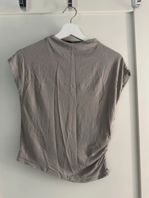 Beige topp med rynkade detaljer - Säljer en beige topp i mjukt material med korta ärmar och hög hals. Toppen har en snygg rynkad detalj på sidan som ger en cool twist till looken. Perfekt att styla med jeans eller kjol för en trendig outfit. Storlek M från lager 157. Säljs då jag har för mycket kläder!