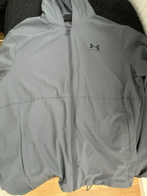 Grå vindjacka från Under Armour - Snygg grå vindjacka från Under Armour med huva och hel dragkedja framtill. Jackan har två sidofickor och meshfoder på insidan för extra komfort. Perfekt för blåsiga dagar och sportiga outfits.Under armour vindbreaker storlek M  Pris 500kr