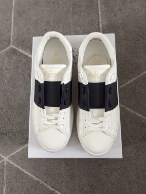 Vita Valentino open - Snygga vita sneakers från Valentino med bred navy rem över snörningen och diskret logga på plösen. Skorna har en chunky sula med rutmönster undertill och är tillverkade i slätt läder. Perfekta för dig som vill ha en clean och exklusiv look.