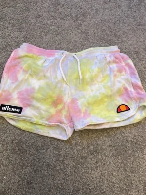 Ellesse pastellfärgade shorts med snörning - Säljer ett par mjuka och bekväma shorts från Ellesse i pastellfärgad tie-dye med inslag av gult, rosa och lite grönt. Shortsen har snörning i midjan, fickor och Ellesse-logga både fram och bak. Perfekta för chill dagar eller träning.