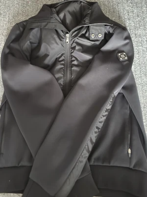 Svart cardigan från j.lindeberg - Snygg svart cardigan med hög krage och dragkedja framtill. Jackan har en blankare yta upptill och matta partier på ärmar och nedtill. Detaljer som bröstpatch och fickor med dragkedja ger en sportig look.
