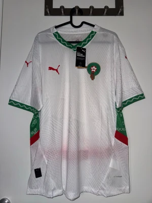 Marocko fotbollströja Puma vit/grön - Snygg officiell Marocko fotbollströja från Puma i vitt med gröna och röda detaljer. Tröjan har korta ärmar, rund hals och ett subtilt mönster i tyget. Marockos landslagsemblem på bröstet och Puma-logga i rött. Tillverkad i lätt och ventilerande material. Ny med lappar.