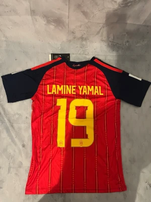 Spaniens landslagströja nr 19 - Snygg röd matchtröja från Spaniens landslag med gula detaljer och nummer 19 samt Lamine Yamal på ryggen. Tröjan har mörkblå ärmar, gula ränder på axlarna och officiella loggor på bröstet. Perfekt för dig som älskar fotboll och vill stötta Spanien.