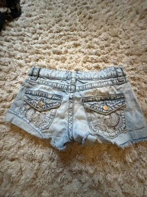 Ljusblå denimshorts med broderi - Säljer ett par ljusblå denimshorts med råa kanter och snygga slitningar. Shortsen har bakfickor med dekorativt broderi och knappar i metall. Perfekta för varma dagar och ger en avslappnad vibe.