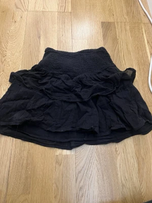 Svart volangkjol från Gina Tricot - Supersöt svart kortkjol från Gina Tricot med bred smock i midjan och flera lager volanger. Kjolen är gjord i ett mjukt och lätt tyg som ger en luftig känsla. Perfekt att styla med en enkel topp eller hoodie.