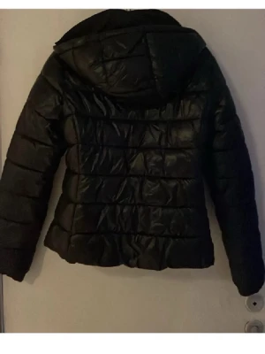 Svart pufferjacka med huva - Snygg svart pufferjacka med huva och dragkedja framtill. Jackan har quiltad design med breda kanaler och två sidofickor med dragkedja. Perfekt för kalla dagar och ger en cool look till vinterns outfits.