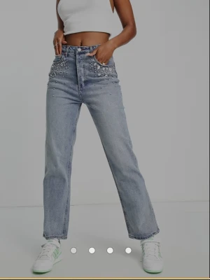 Oanvända stradivarius mom jeans 36/S - Säljer ett par ljusblå raka jeans med hög midja och snygga pärl- och stendekorationer framtill vid fickorna. Jeansen har klassisk femficksdesign och är tillverkade i denim. 100% bomull. Oanvända med tags kvar. Storlek 36/S