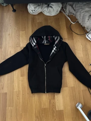 Svart hoodie med dragkedja från Burberry - Snygg svart hoodie från Burberry med dragkedja framtill och klassiskt rutigt foder i huvan. Hoodien har långa ärmar och ribbade muddar. Perfekt för dig som vill ha en stilren och exklusiv look. Materialet känns mjukt och bekvämt.