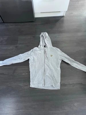 Ljusgrå zip hoodie från Lyle & scott  - Säljer en ljusgrå hoodie med dragkedja och huva. Tröjan har långa ärmar, fickor framtill och en liten gul logga på bröstet. Tillverkad i mjukt bomullsmaterial och har snören i huvan för justering. Perfekt för en avslappnad stil.