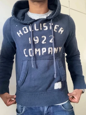 Mörkblå hoodie Hollister 1922 - Mörkblå hoodie från Hollister med trycket 'Hollister 1922 Company' på bröstet. Klassisk känguruficka framtill, dragsko i huvan och ribbade muddar. Mjuk bomullsmix som känns skön mot huden. Perfekt för en avslappnad och cool stil. 