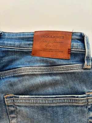 Blå jeans från Jack & Jones - Säljer ett par klassiska blå jeans från Jack & Jones med snygg tvätt och raka ben. Jeansen har fem fickor, bälteshällor och detaljer i kontrastsömmar. Materialet är slitstarkt denim och insidan har ett coolt blommigt tygmönster.