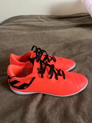 Adidas orange fotbollsskor 19.4 - Säljer ett par snygga orange fotbollsskor från Adidas modell 19.4 med svarta snören och detaljer. Skorna har låg profil och är tillverkade i syntetmaterial med vit sula, perfekta för inomhusspel. Stilren design med Adidas-logga på plösen.