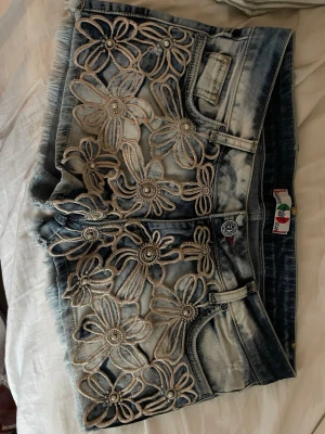 Blå jeansshorts med blommig spets - Säljer ett par ljusblå jeansshorts med råa kanter och stora blommiga spetsdetaljer framtill. Shortsen har fem fickor, knappgylf och cool tvättad look. Perfekta för sommaren och ger en unik touch till din outfit!