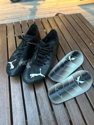 Svarta Puma Future fotbollsskor - Säljer ett par svarta Puma Future fotbollsskor med vita detaljer och klassisk logga på tån. Skorna har snörning och medföljer två svarta/vita benskydd. Snygg och modern design, perfekt för dig som vill sticka ut på planen.