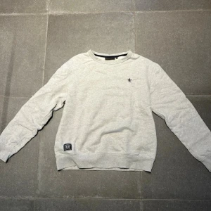 Ljusgrå sweatshirt från Morris - Säljer en ljusgrå sweatshirt från Morris med rund halsringning och diskret logga broderad på bröstet. Tröjan har ribbade muddar vid ärmslut och nederkant samt en liten patch nertill. Perfekt för en avslappnad stil.