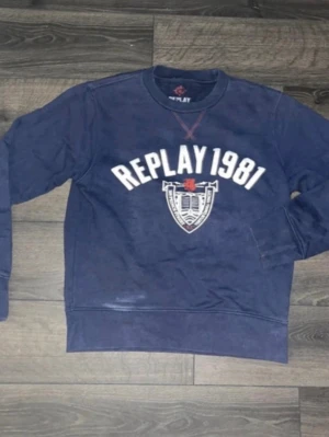 Replay tröja  - Säljer en marinblå sweatshirt från Replay med trycket 'REPLAY 1981' och ett coolt emblem framtill. Tröjan har rund hals, långa ärmar och ribbade muddar. Perfekt för dig som gillar klassisk streetstyle och vill ha något bekvämt och snyggt. Som det syns har tröjan en blekning/fläck långt ned till vänster på framsidan.OBS! Tröjan har möjligen krympt i tvätten så kolla måtten noggrant.