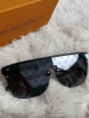 Svarta Louis Vuitton solglasögon - Snygga svarta solglasögon från Louis Vuitton med breda, rektangulära glas och spegeleffekt. Glasen har diskret LV-mönster och bågarna är i svart. Perfekt accessoar för att höja din stil.