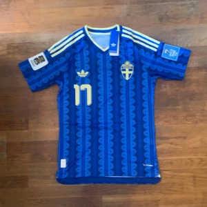 Sverige VM tröja Gyökeres #17 - Säljer en blå fotbollströja från Adidas med Sveriges landslagsemblem och nummer 17. Tröjan har gula detaljer, vita ränder på axlarna och namnet Gyökeres på ryggen. Tillverkad i lätt och ventilerande material, perfekt för match eller träning.