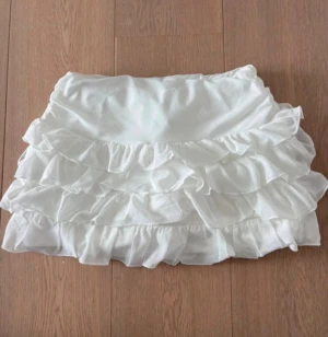 Vit volangskort kjolshorts - Supersöta vita skorts med flera lager volanger. Kjolshortsen har en elastisk midja och är gjorda i ett mjukt, lätt tyg som ger en flowy känsla. Perfekt för dig som gillar en feminin och trendig stil med extra rörelse i plagget.