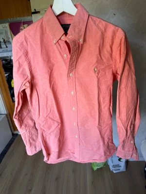 Rosa/korall skjorta från Polo Ralph Lauren - Säljer en snygg rosa/korall skjorta från Polo Ralph Lauren med klassisk krage och knappar framtill. Skjortan har lång ärm och den ikoniska broderade loggan på bröstet. Perfekt för dig som vill ha en stilren och tidlös look.