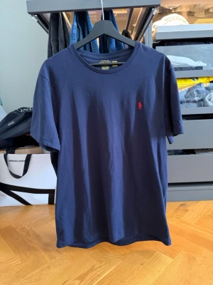 Mörkblå Ralph lauren t-shirt - Snygg mörkblå Ralph lauren t-shirt i bomull med klassisk rund hals och korta ärmar. På bröstet finns en liten röd broderad logga som ger en stilren touch. Perfekt basplagg för en enkel och clean look. STOR I HALSEN! 