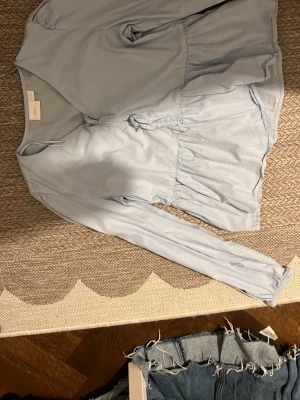 Ljusblå blus med volang - Snygg ljusblå blus med volangdetaljer i midjan och långa ärmar. Blusen har en avslappnad passform och knytning framtill. Perfekt för en trendig och stilren look.