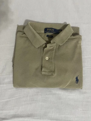 Beige pikétröja från Polo Ralph Lauren - Klassisk beige pikétröja från Polo Ralph Lauren med korta ärmar och krage. Tröjan har två knappar framtill och den ikoniska mörkblå logotypen broderad på bröstet. Perfekt till jeans eller shorts för en clean och stilren look. 