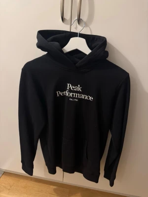 Svart Peak Performance hoodie - Snygg svart hoodie från Peak Performance med vit logga på bröstet. Huva och ficka framtill. Passar XS-S