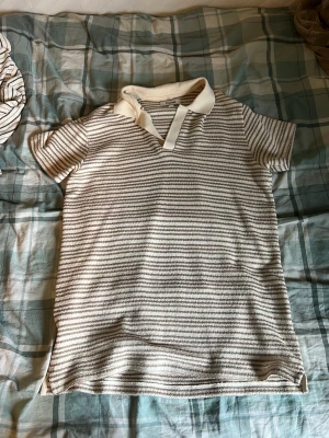 Randig t-shirt med krage - Säljer en vit och grå randig t-shirt med korta ärmar och klassisk krage. T-shirten har en v-ringad öppning framtill och är gjord i ett mjukt bomullsliknande material. Perfekt för en avslappnad och stilren look.