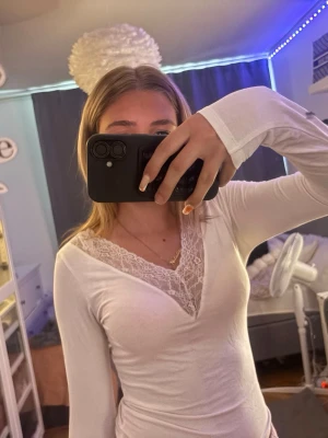Vit långärmad topp med spets - Snygg vit långärmad topp med v-ringning och spetsdetaljer vid halsen. Toppen har en slim passform och är perfekt för dig som gillar en enkel men ändå feminin stil. Tillverkad i mjukt material som sitter skönt på kroppen.