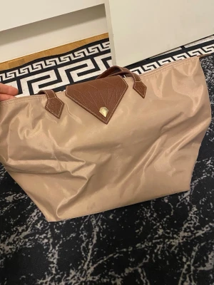 Beige shoppingväska med bruna detaljer - Stilren beige shoppingväska med bruna handtag och lockdetalj. Väskan har en enkel, rymlig design och är perfekt för att bära allt du behöver. Materialet är troligtvis syntet med detaljer i skinnimitation. Passar till alla dina ärenden i stan.