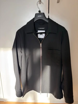 Svart overshirt från J.Lindeberg - Stilren svart overshirt från J.Lindeberg med rak passform, klassisk krage och silverfärgad dragkedja framtill. En bröstficka på vänster sida och långa ärmar med knapp vid ärmslut. Perfekt lager på lager-plagg för en clean look.