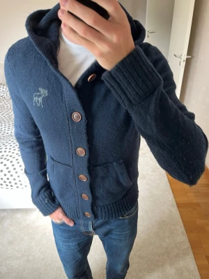 Mörkblå kofta med huva och knappar från Abercrombie&Fitch - Säljer en sjukt snygg och unik stickad hoodie från Abercrombie & Fitch som är i ett fint skick (lite nopprig på armbågen men går att ta bort med noppermaskin enkelt då den blir som ny!). ———————————————————                                                📐| Storlek: M                                                             👖| Märke: Abercrombie & Fitch                                      🔎| Skick: Fint skick!                                              ✍🏻| Skriv om du har några frågor