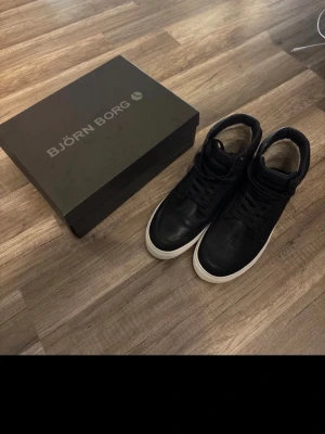 Svarta Björn Borg sneakers, storlek 43 - Snygga svarta sneakers från Björn Borg i storlek 43. Skorna har en klassisk design med högt skaft, svarta skosnören och vit sula. Tillverkade i ett material som ser ut som läder, perfekta för en clean och stilren look.